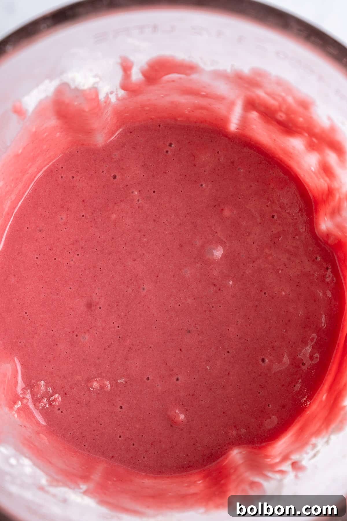 A clear glass is filled with a thick, velvety pink smoothie, exhibiting a creamy consistency and delicate small bubbles on its uneven surface. The vibrant color suggests a rich blend of fruits, such as berries, creating an inviting and refreshing visual appeal.
