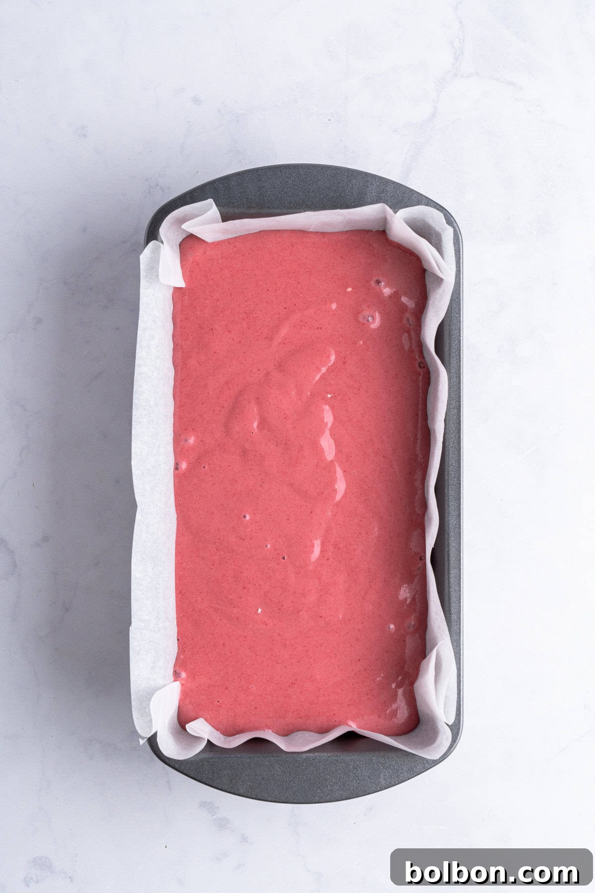 A loaf pan, neatly lined with parchment paper, is filled to the brim with a smooth, pink batter, poised and ready for baking. The pan rests gracefully on a pristine light marble surface, anticipating its transformation in the oven.