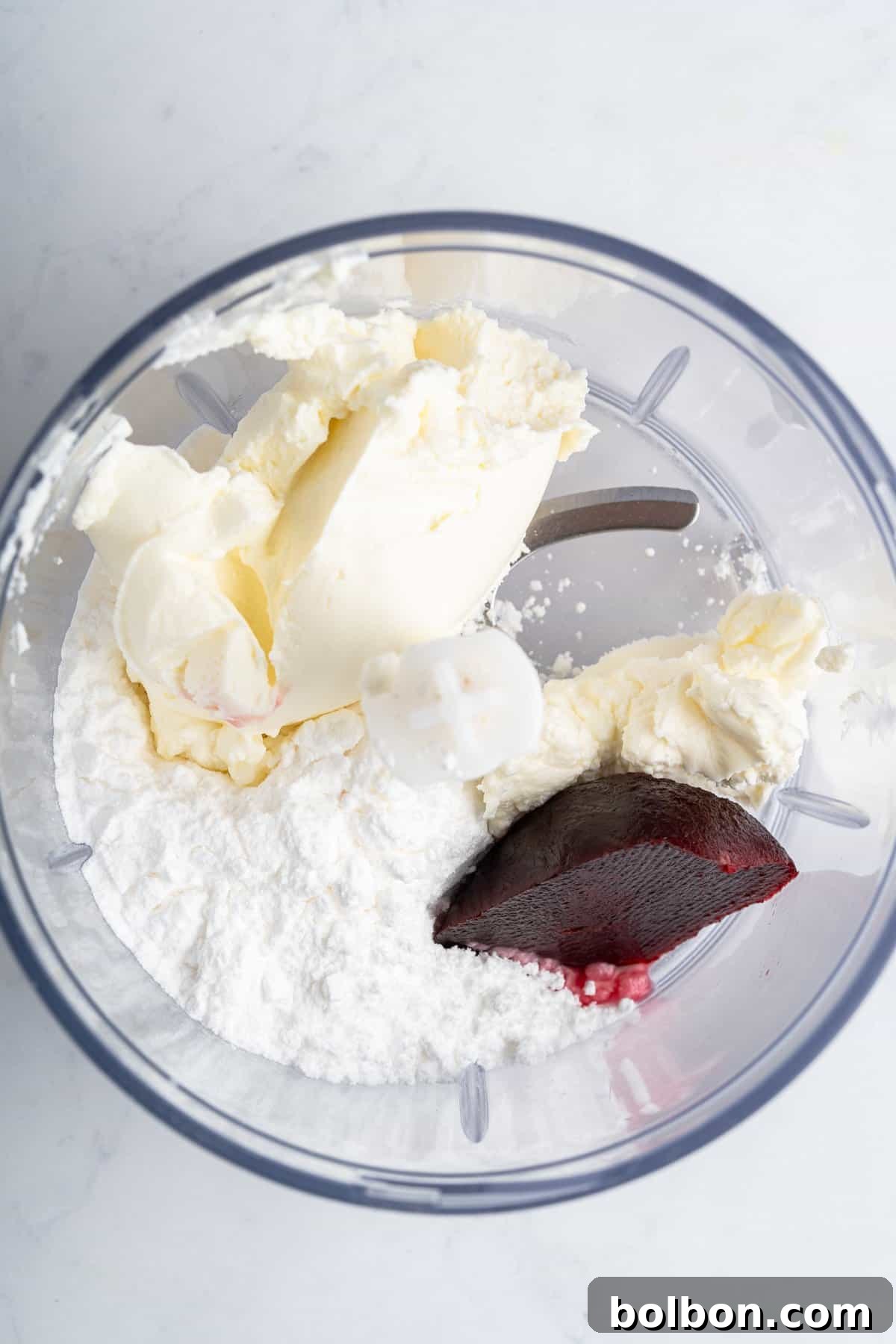 Inside a food processor bowl, cream cheese, powdered sugar, and a vibrant dollop of beet puree (or red food coloring) are ready for blending. The ingredients are neatly arranged on a clean, light countertop, awaiting their transformation into a smooth, colorful frosting.