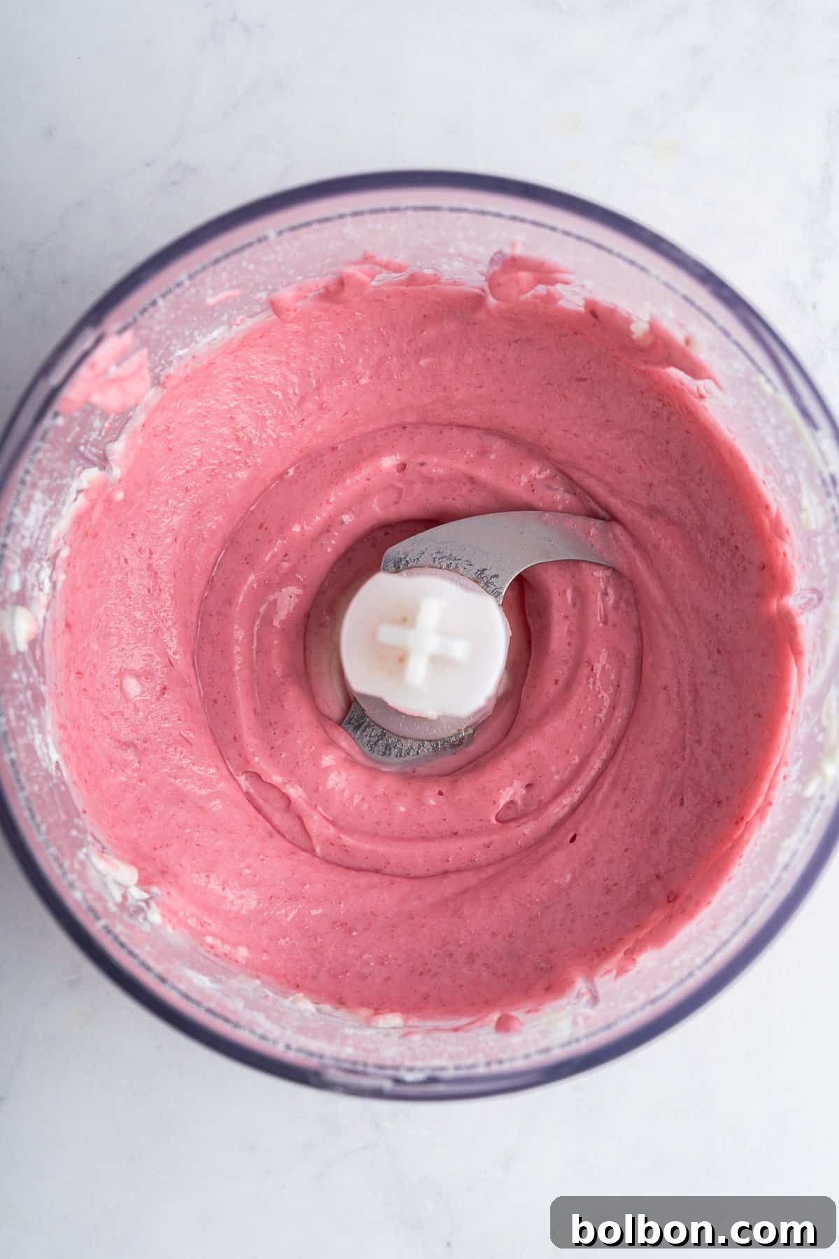 A top-down perspective captures the smooth, uniformly blended pink mixture resting inside a food processor bowl. The metal blade is clearly visible in the center, nestled within the creamy consistency of the mixture. The backdrop is a subtly textured white surface.