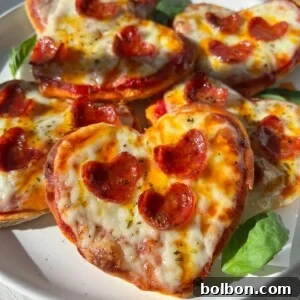 A plate of delightful Heart Shaped Pizza minis topped with melted cheese, small pepperoni slices, and fresh basil leaves sits on a white plate, basking in bright sunlight.
