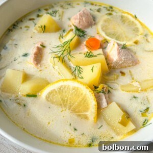 A bowl of creamy Finnish Salmon Soup with chunks of salmon, potatoes, carrot slices, lemon wedges, and fresh dill, served in a white bowl on a light wooden surface.