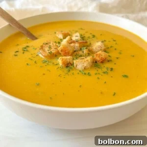 A bowl of creamy orange Cream of Zucchini Soup topped with croutons and sprinkled herbs, with a spoon resting inside.