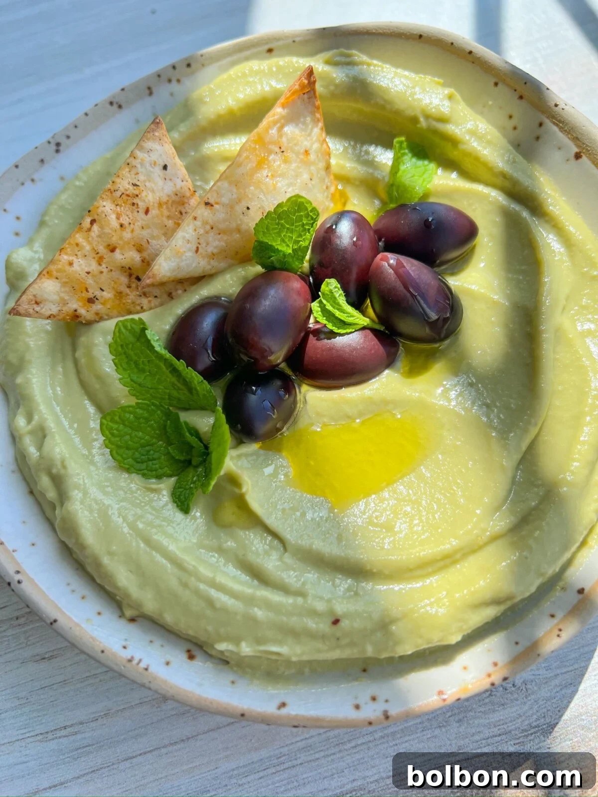 Avocado Hummus in a light bowl, garnished with olives, mint leaves, and olive oil, with a subtle background of seasoned tortilla chips.