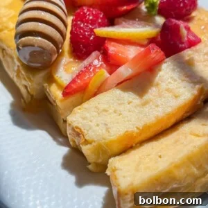 A close-up of fluffy, sliced French toast topped with fresh strawberries, raspberries, lemon slices, and drizzled with honey from a honey dipper, served on a white plate alongside warm No Knead Garlic Bread in bright natural light.