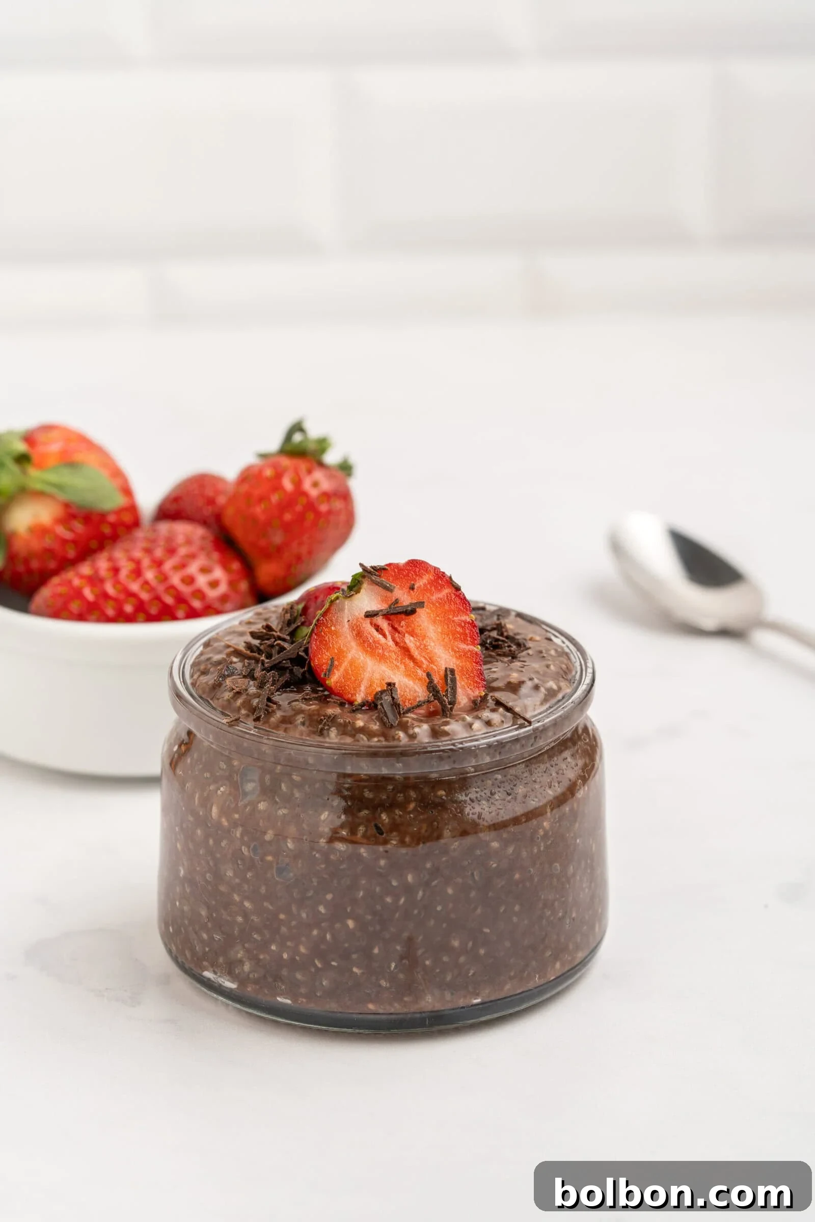 A glass jar of chocolate chia pudding topped with sliced strawberries and chocolate shavings sits on a white surface. In the background, there is a bowl of fresh strawberries and a spoon.