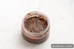 A small glass jar filled with a thick, dark brown, slightly textured mixture sits on a white, lightly marbled surface.
