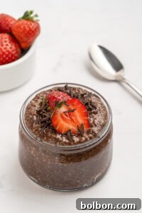 A glass jar filled with chocolate chia pudding, topped with a sliced strawberry and chocolate shavings, sits on a white surface with a spoon and a bowl of whole strawberries in the background.