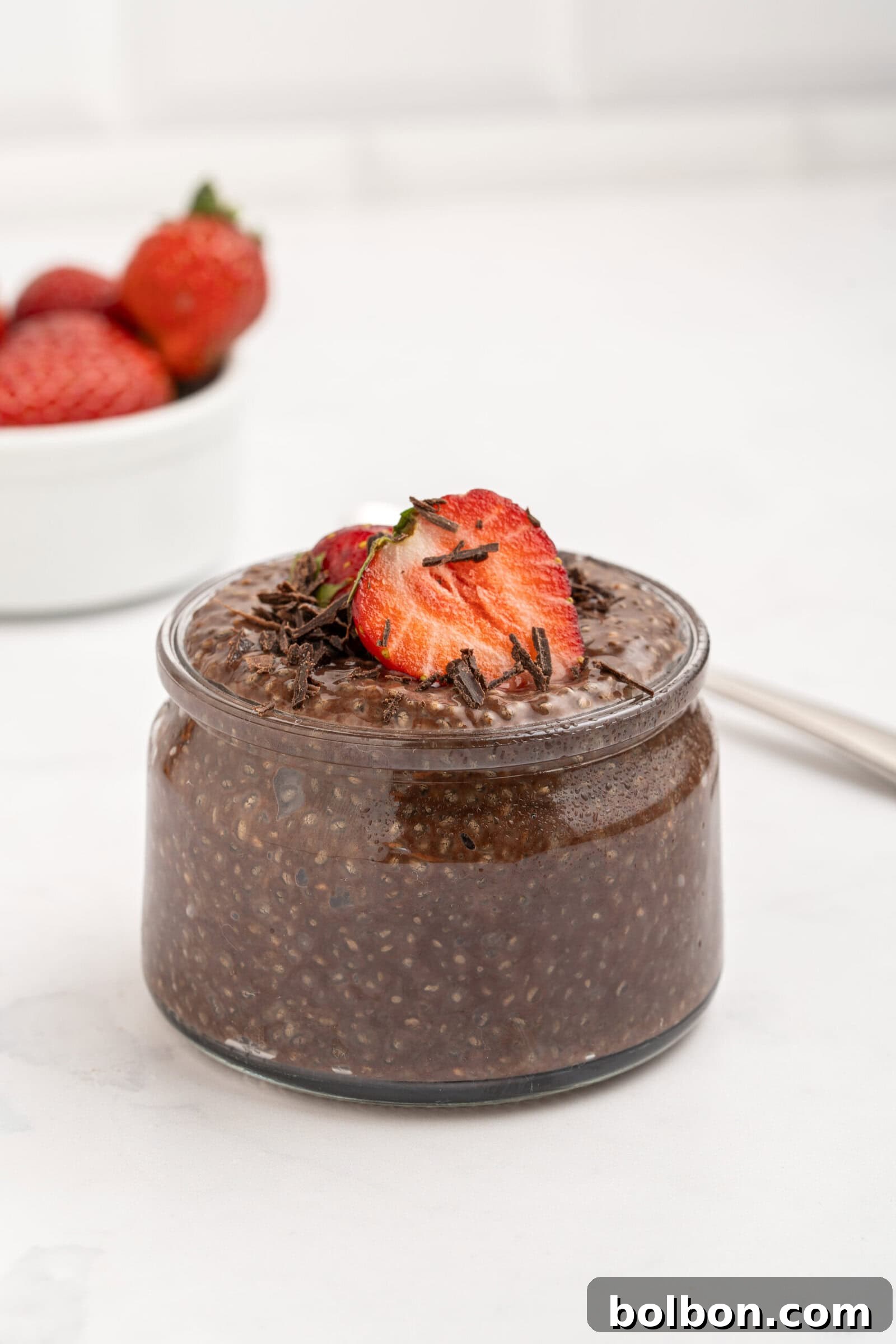 A small glass jar filled with chocolate chia pudding, topped with sliced strawberries and chocolate shavings. In the background, a white bowl of whole strawberries sits on a white surface.