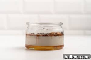 A glass jar on a white surface contains separated layers of honey, milk, cinnamon, and vanilla, against a white tiled background.