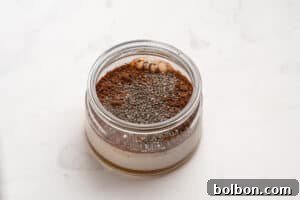A glass jar filled with layers of milk, cocoa powder, chia seeds, and nut butter sits on a white marble surface.