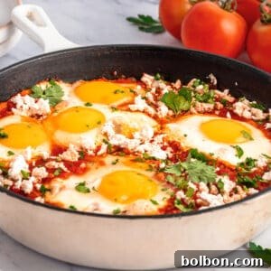 A skillet filled with a classic Shakshouka Recipe, featuring poached eggs in tomato sauce, topped with crumbled cheese and chopped parsley. Fresh tomatoes, parsley, and slices of bread are in the background.