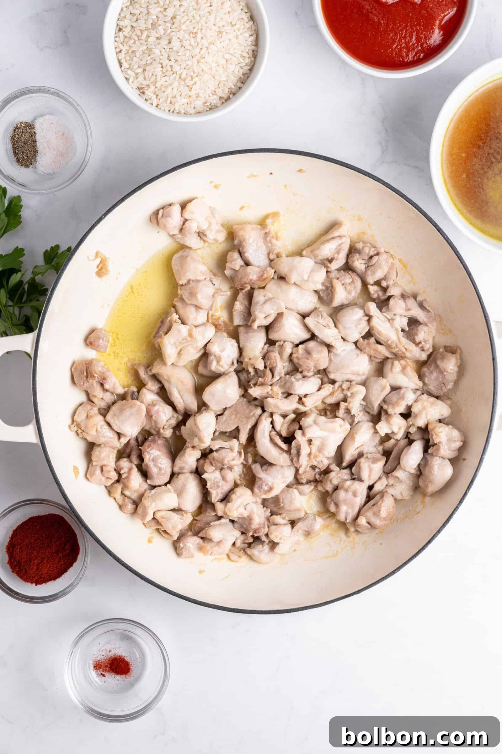A pan of sautéed chicken pieces on a white surface, surrounded by bowls containing rice, tomato sauce, broth, spices, and fresh parsley.