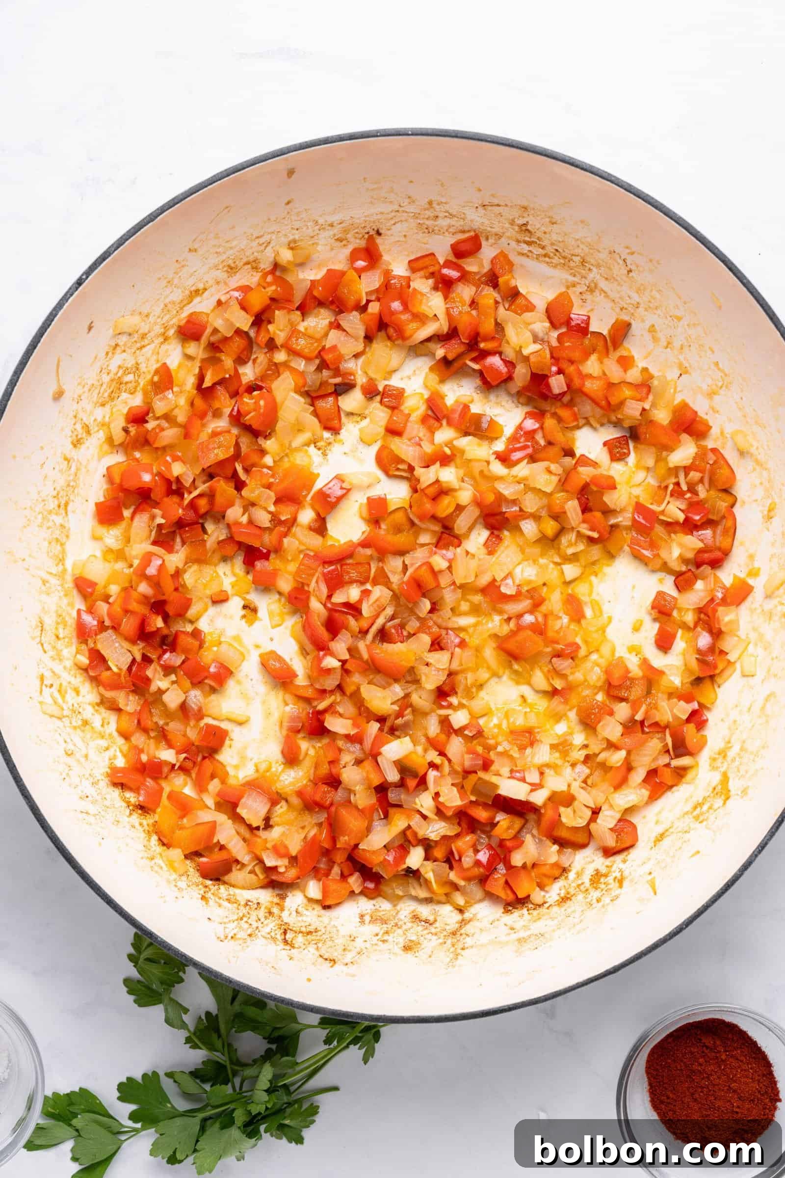 Diced red peppers and onions sautéed in a large pan on a white surface. Fresh parsley and a small bowl of paprika are nearby.
