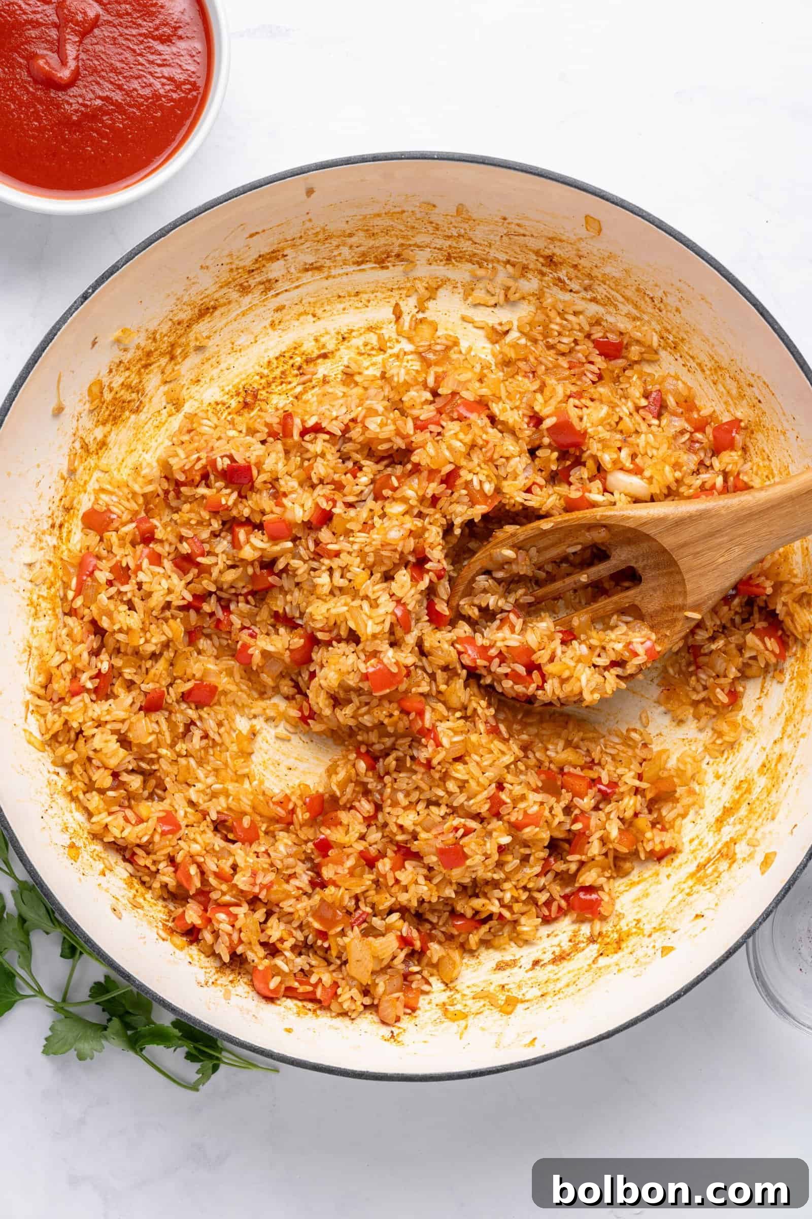 A pot of seasoned rice mixed with chopped red peppers and onions. A wooden spoon rests in the rice. A bowl of tomato sauce is visible in the top left corner. Fresh parsley is partially visible at the bottom left.