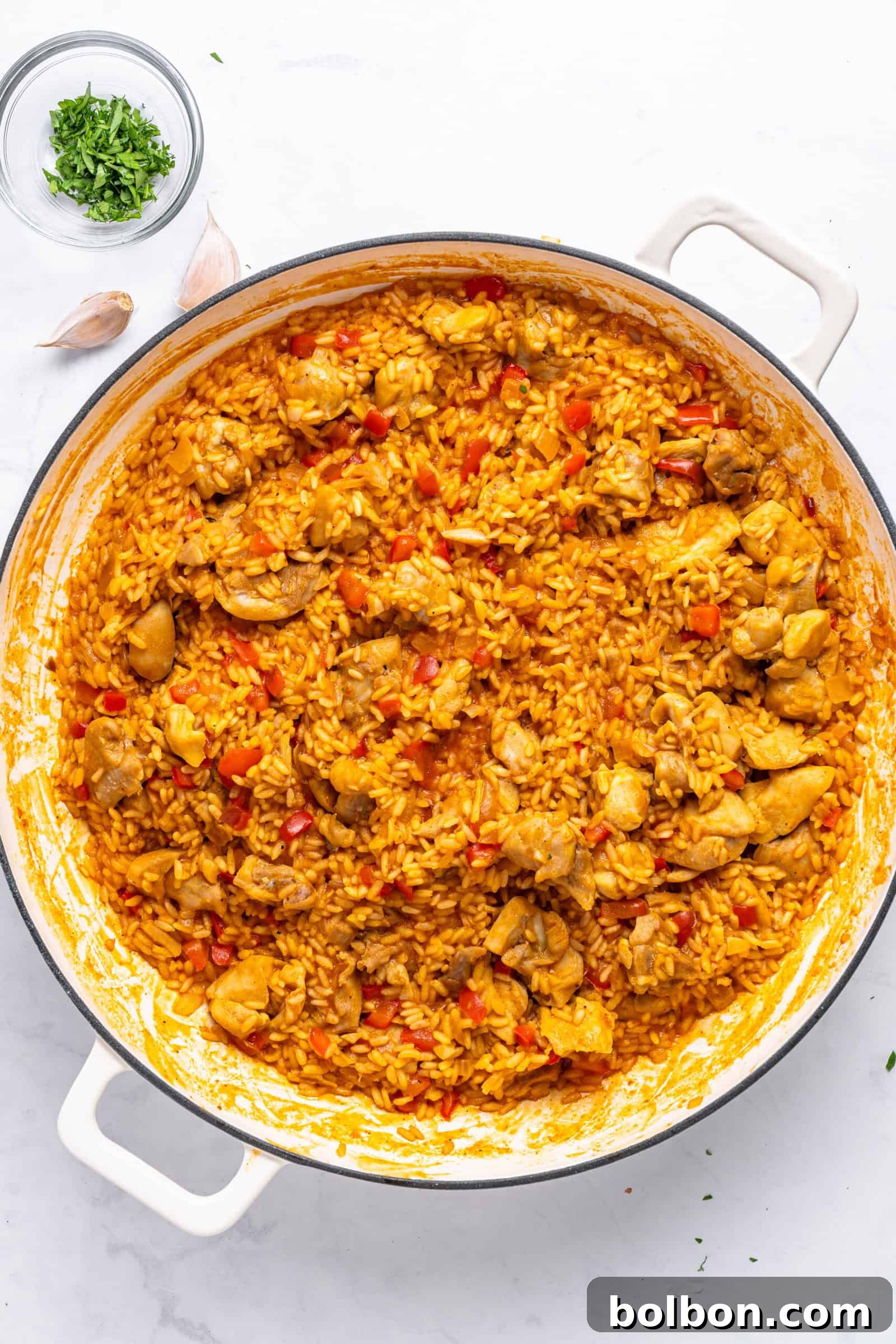 A large pot of vibrant yellow rice mixed with chunks of chicken and red bell peppers, garnished with fresh herbs. It is set on a white marble surface with scattered garlic cloves and a small bowl of chopped herbs nearby.