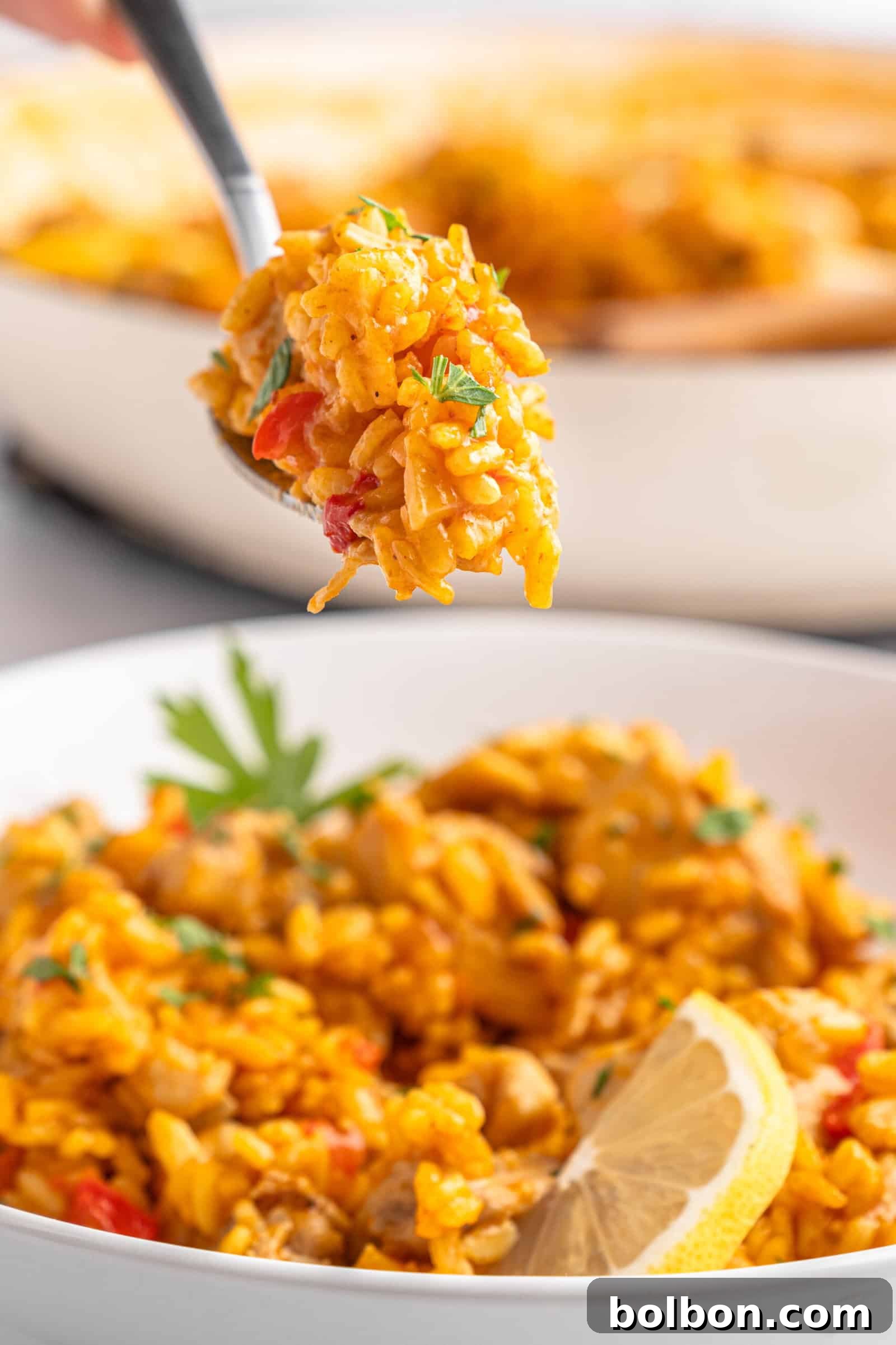 A close-up of a spoon holding a portion of rice over a bowl filled with the dish. The plate features yellow rice, diced vegetables, and garnished with parsley and a lemon wedge. 