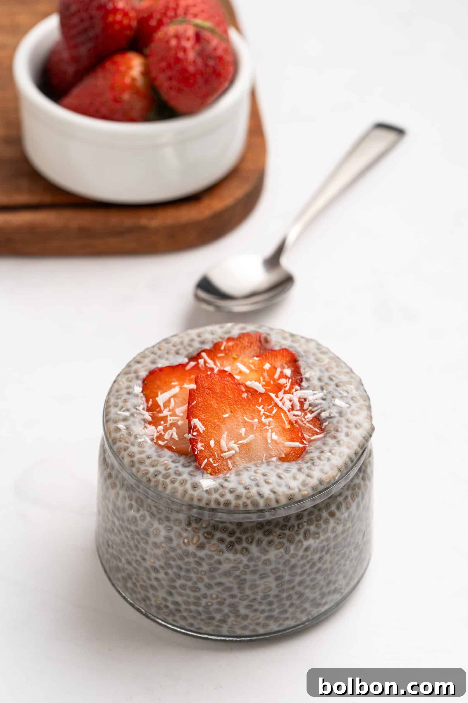 Wholesome Chia Delight 2 A glass jar of chia pudding topped with sliced strawberries and shredded coconut, with a spoon beside it and a bowl of whole strawberries in the background.