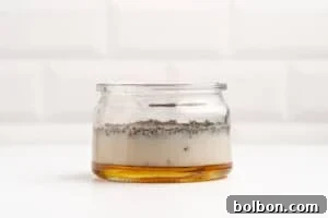 A glass jar containing separated layers of honey, a creamy substance, and chia seeds, set against a white tiled background.