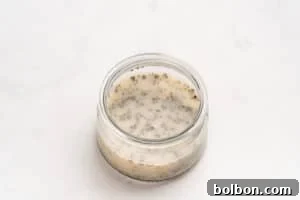 A small glass jar filled with a light-colored mixture containing chia seeds, sitting on a white, lightly textured surface.