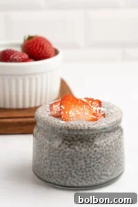 A small glass jar filled with chia pudding, topped with sliced strawberries and shredded coconut. In the background, a white bowl holds more strawberries on a wooden tray.