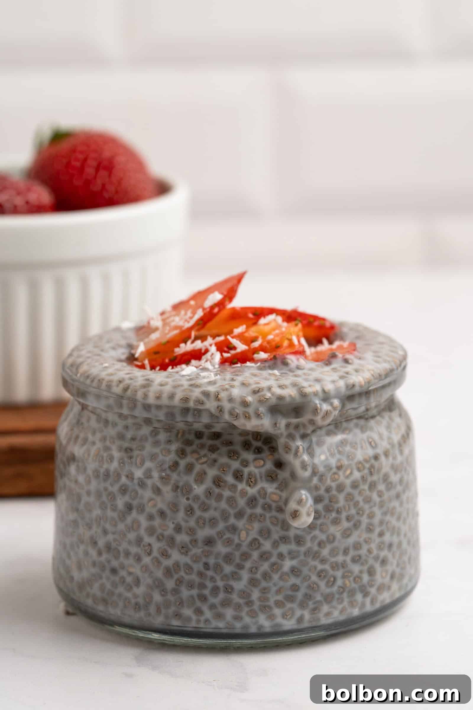 Wholesome Chia Delight 4 A glass jar filled with chia pudding is topped with sliced strawberries and shredded coconut. In the background, a white bowl holds whole strawberries. The scene is set on a white surface with a white tiled wall.