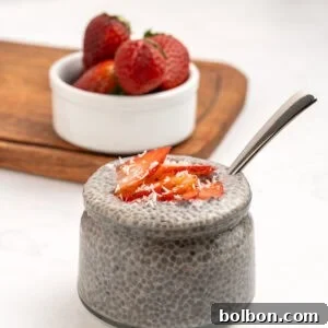 A glass jar filled with chia pudding, topped with sliced strawberries and shredded coconut, sits on a white surface. A spoon is inside the jar, and a small bowl of whole strawberries is in the background on a wooden board.
