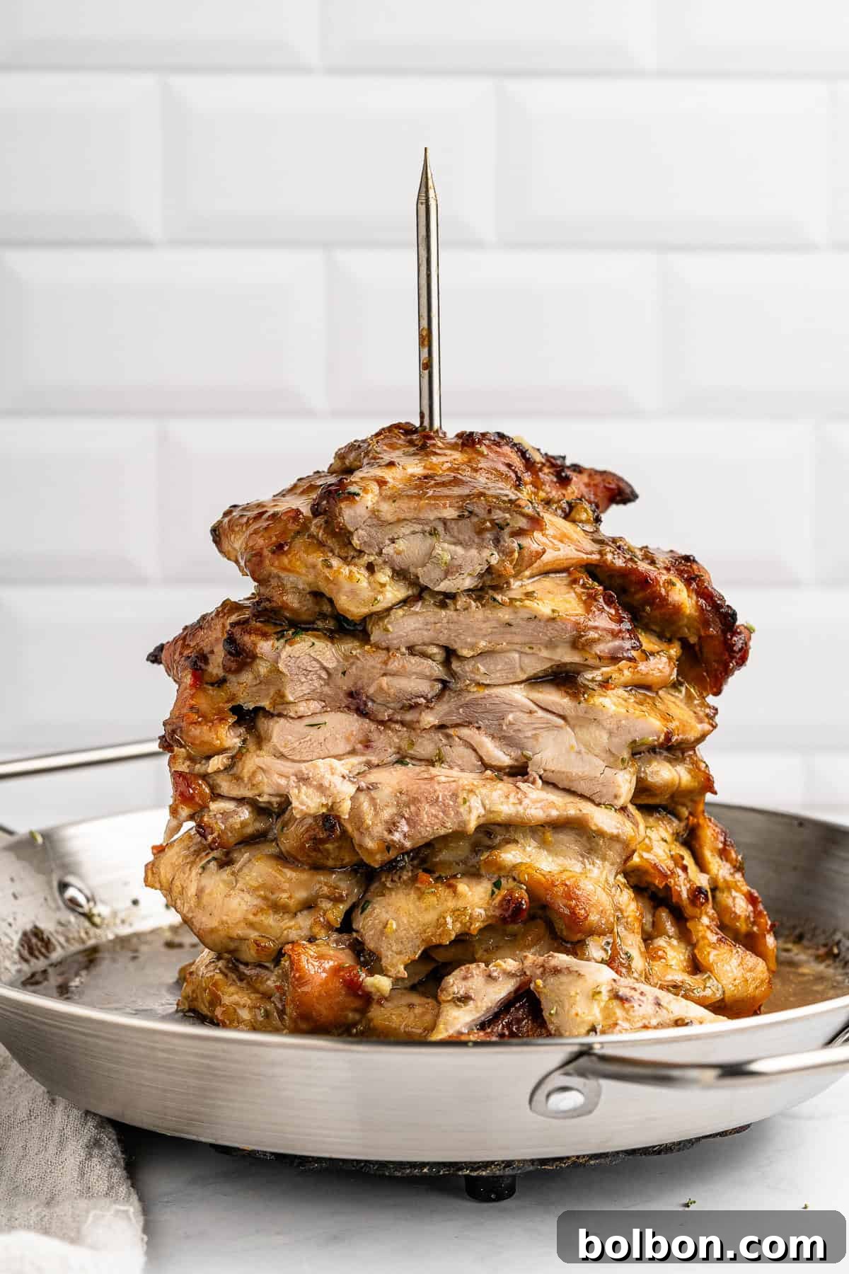 Homemade Greek Chicken Gyros 3 Stacked layers of marinated, grilled meat on a vertical skewer in a metal pan, set against a white tiled background.