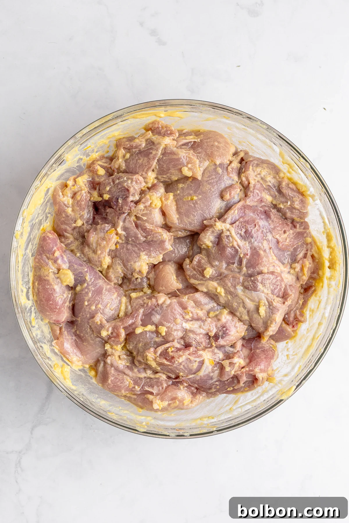Homemade Greek Chicken Gyros 6 A bowl of raw marinated chicken pieces in a light yellow marinade, placed on a white marble surface. The marinade appears to contain visible chunks of seasoning or spices.
