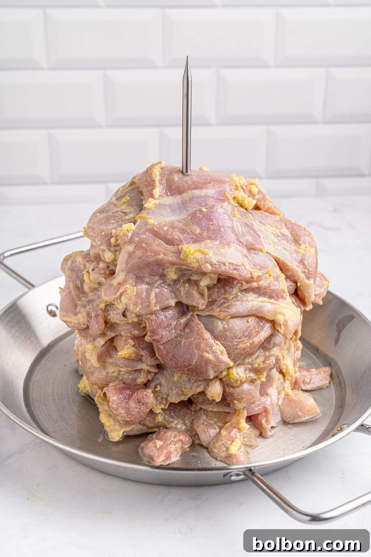 Homemade Greek Chicken Gyros 7 Raw marinated meat stacked on a skewer, placed vertically on a metal pan, ready for grilling. The background features white tiled walls.