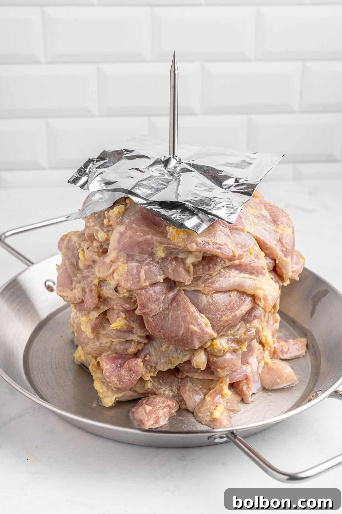 Homemade Greek Chicken Gyros 8 Raw marinated meat stacked vertically on a skewer in a metal tray, with a foil sheet covering the top. The background features white tiled walls.