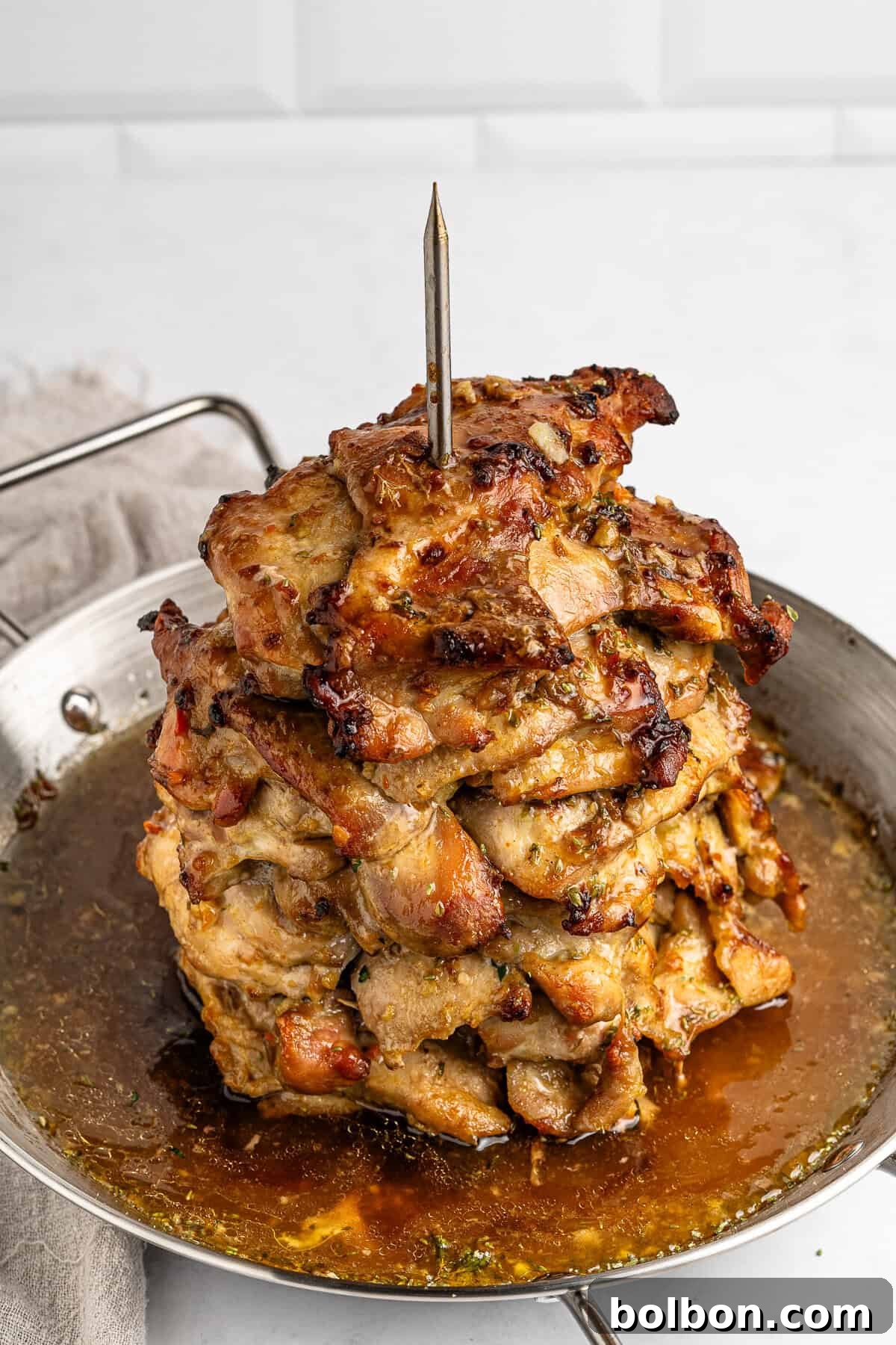 Homemade Greek Chicken Gyros 9 A stack of marinated and grilled meat pieces is skewered vertically on a spit in a round metal pan. The meat is browned and juicy, surrounded by flavorful juices. A beige cloth napkin is partially visible on the left side.