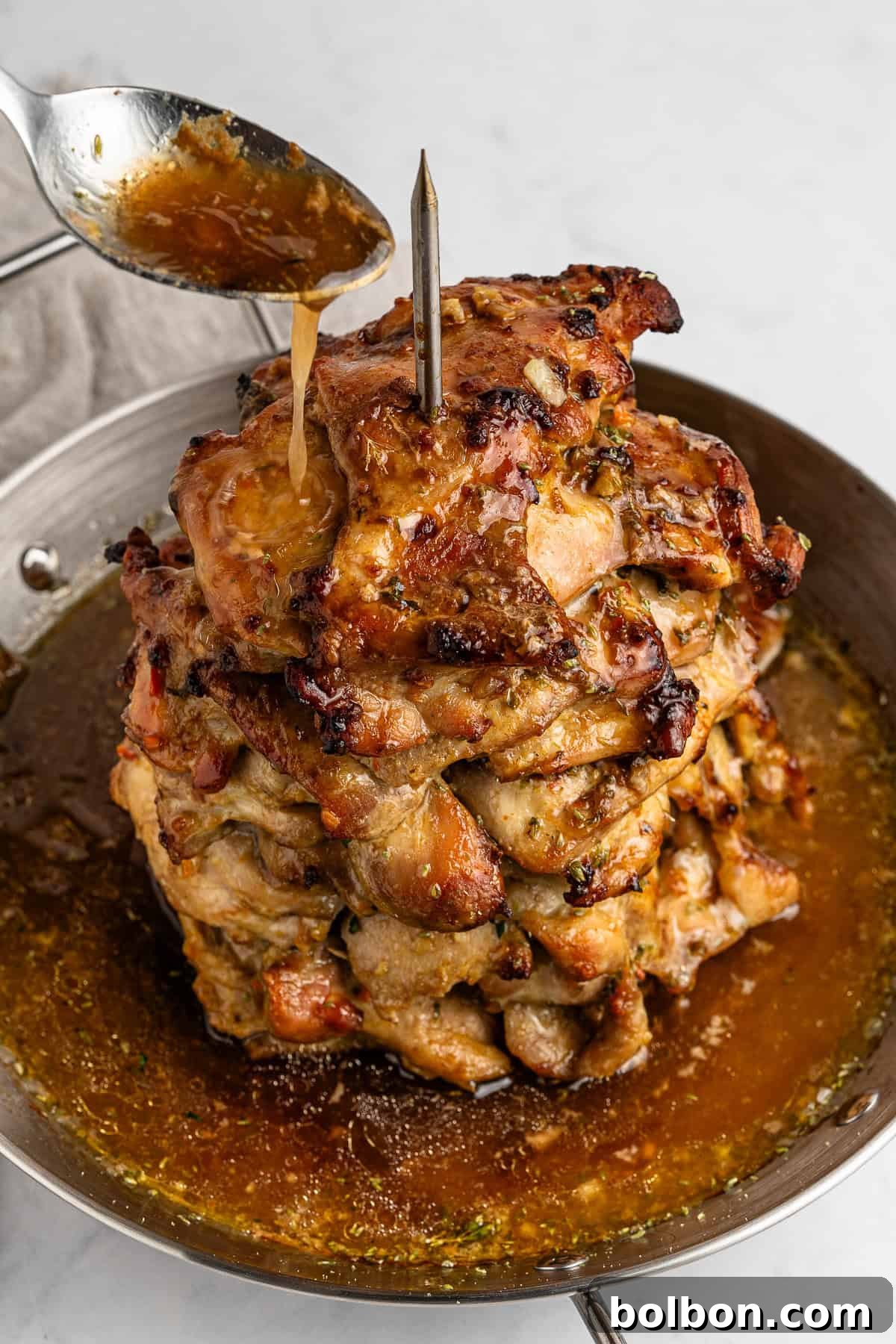 Homemade Greek Chicken Gyros 10 A vertical stack of marinated, grilled meat on a skewer in a pan, with a spoon drizzling sauce over it. The meat is browned and juicy, sitting in a pool of sauce. The background is a light, neutral countertop.