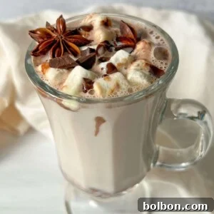 A glass mug filled with creamy hot chocolate made from Kinder Hot Chocolate Bombs, topped with marshmallows, chocolate shavings, and star anise, sits on a white surface with a light-colored cloth in the background.