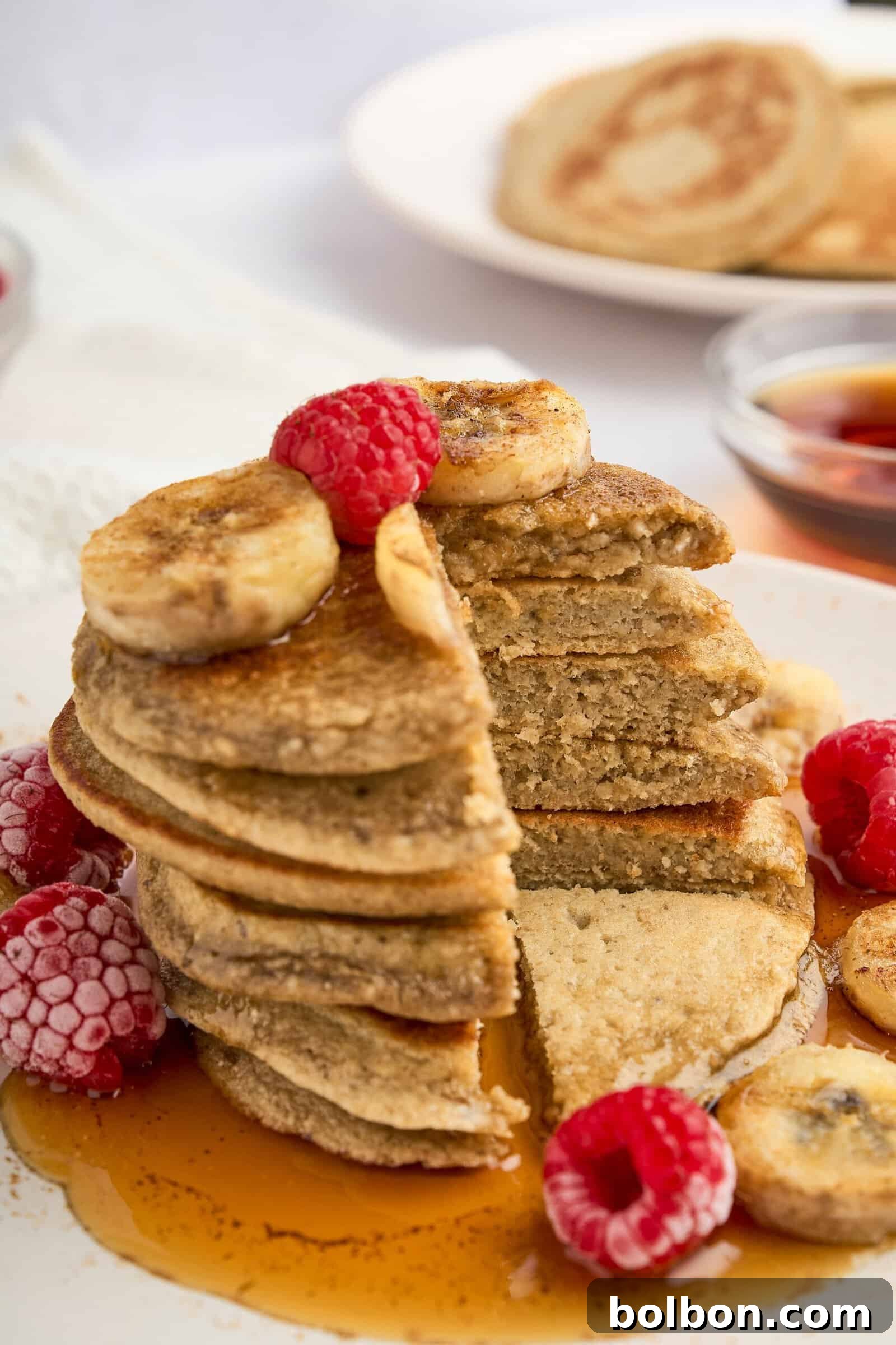 Guilt Free Banana Pancakes 2 A stack of healthy banana pancakes with a slice cut out, topped with banana slices, raspberries, and drizzled with syrup. Raspberries and syrup are also on the plate. Another plate with pancakes is blurred in the background.