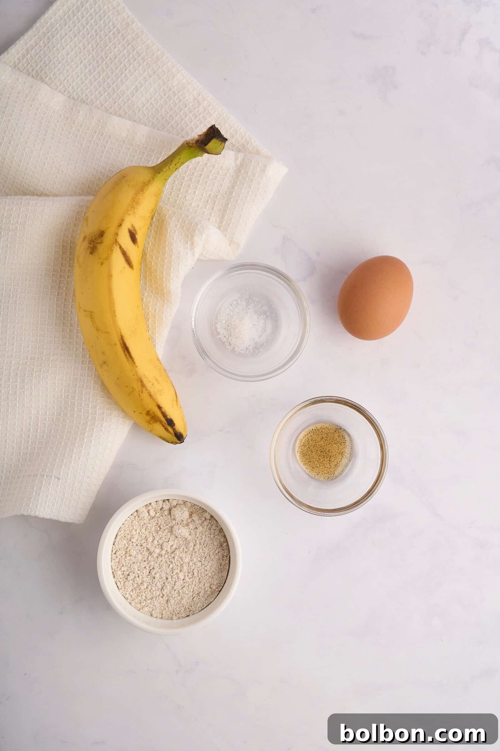 Guilt Free Banana Pancakes 3 Top view of a banana, an egg, a small bowl of flour, a bowl of white powder (possibly sugar or salt), and a bowl of ground spices on a light surface with a textured cloth. Ingredients suggest preparation for baking or cooking.