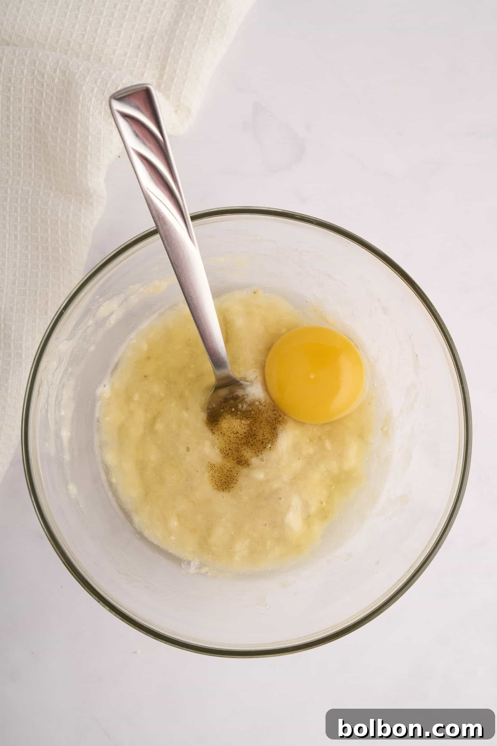 Guilt Free Banana Pancakes 4 A glass bowl with mashed bananas, a cracked egg, and vanilla extract. A metal spoon rests in the mixture. A white kitchen towel is in the background, set on a marble countertop.