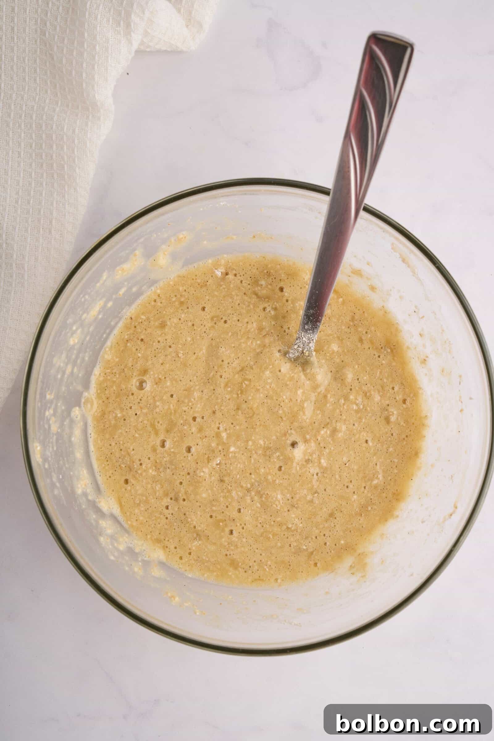Guilt Free Banana Pancakes 5 A glass bowl filled with a light brown cake batter and a silver spoon resting in it. The bowl is placed on a white marble countertop, and a white cloth is partially visible in the top left corner.