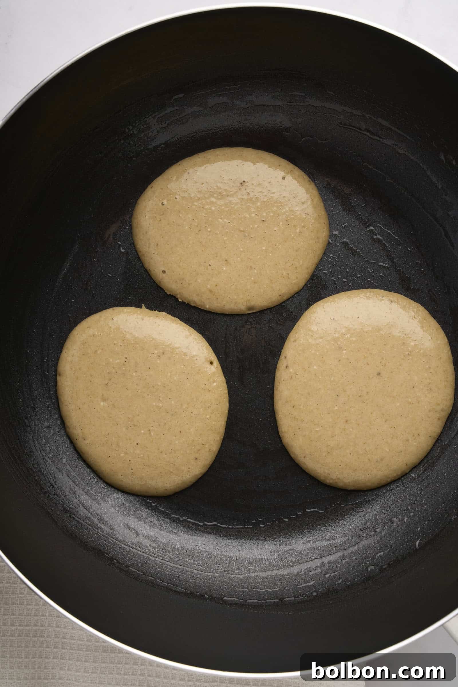 Guilt Free Banana Pancakes 6 Three round pancakes cooking in a non-stick frying pan on a stovetop. The batter is slightly bubbly, indicating it's being cooked on one side. The pan is black with a smooth surface.