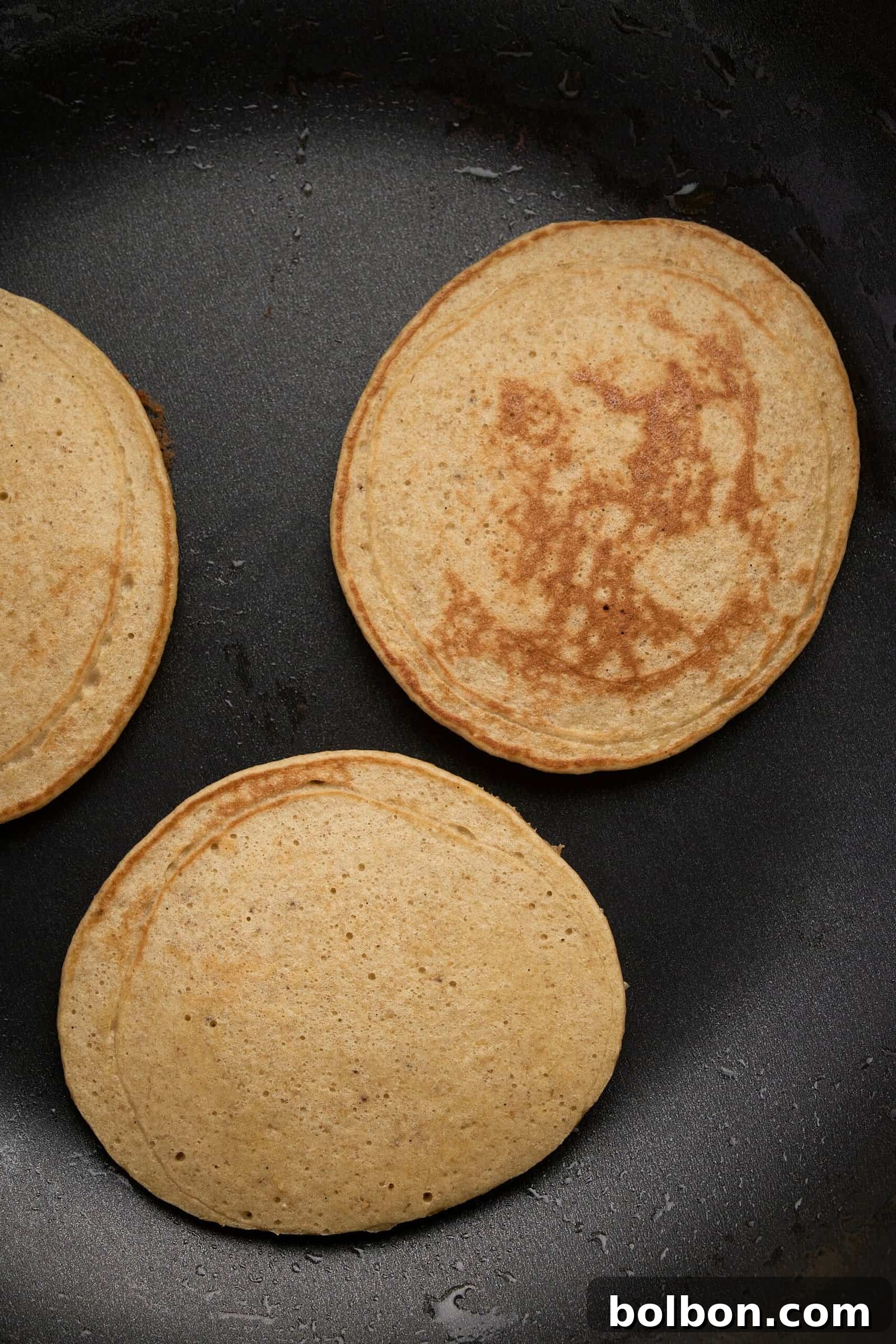 Guilt Free Banana Pancakes 7 Three golden-brown pancakes cooking in a black skillet. The pancakes have small dark spots and are evenly cooked, indicating they are nearing readiness. The skillet surface is smooth and slightly shiny.