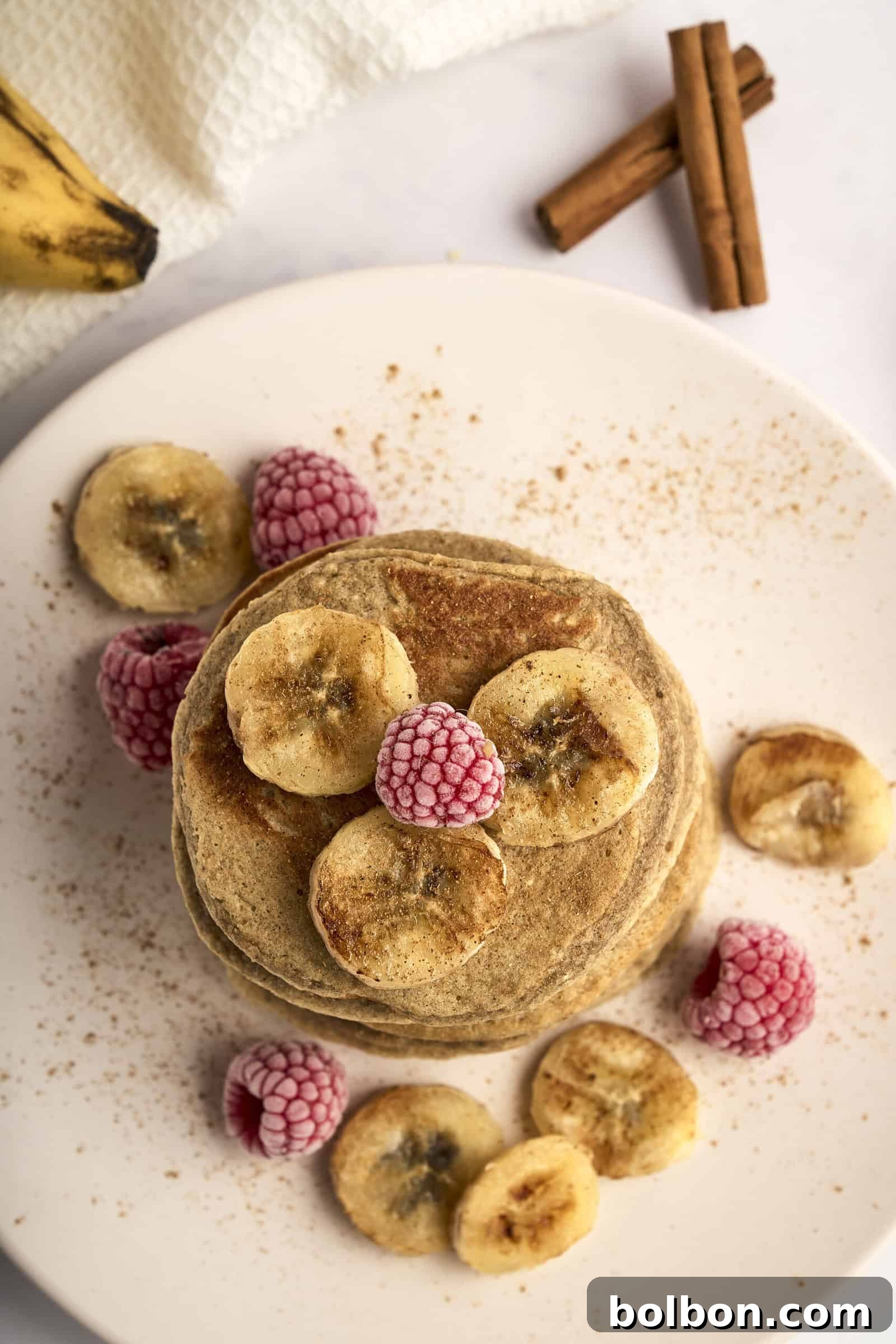 Guilt Free Banana Pancakes 8 A stack of pancakes topped with banana slices and frozen raspberries on a white plate. Cinnamon sticks are in the background, with a banana and a white cloth nearby. The scene conveys a warm, cozy breakfast setting.