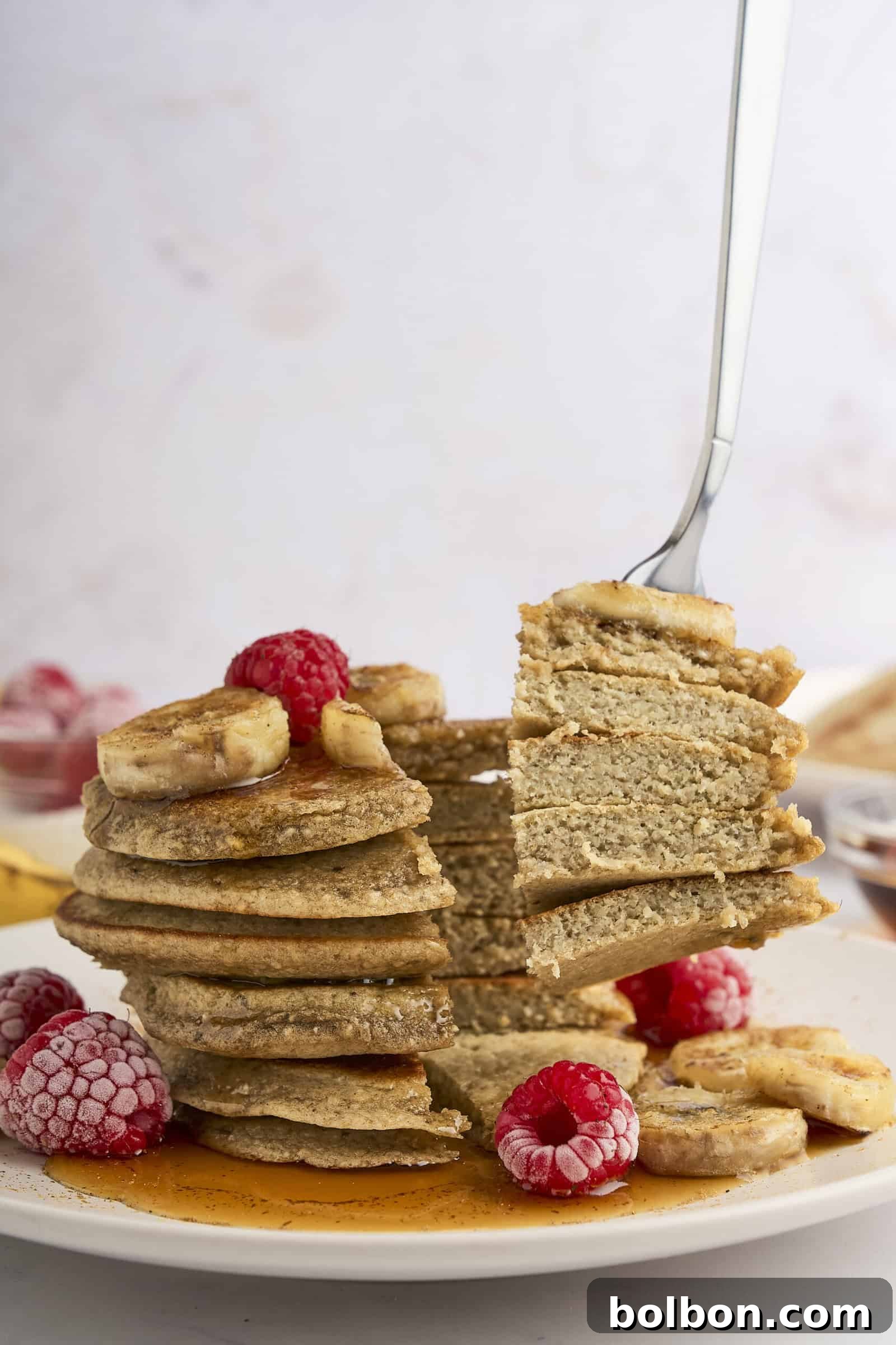 Guilt Free Banana Pancakes 9 A stack of healthy banana pancakes topped with banana slices, raspberries, and syrup. A fork is lifting a section of the pancakes, showing the fluffy texture. Some frozen raspberries and banana slices are scattered around the plate.