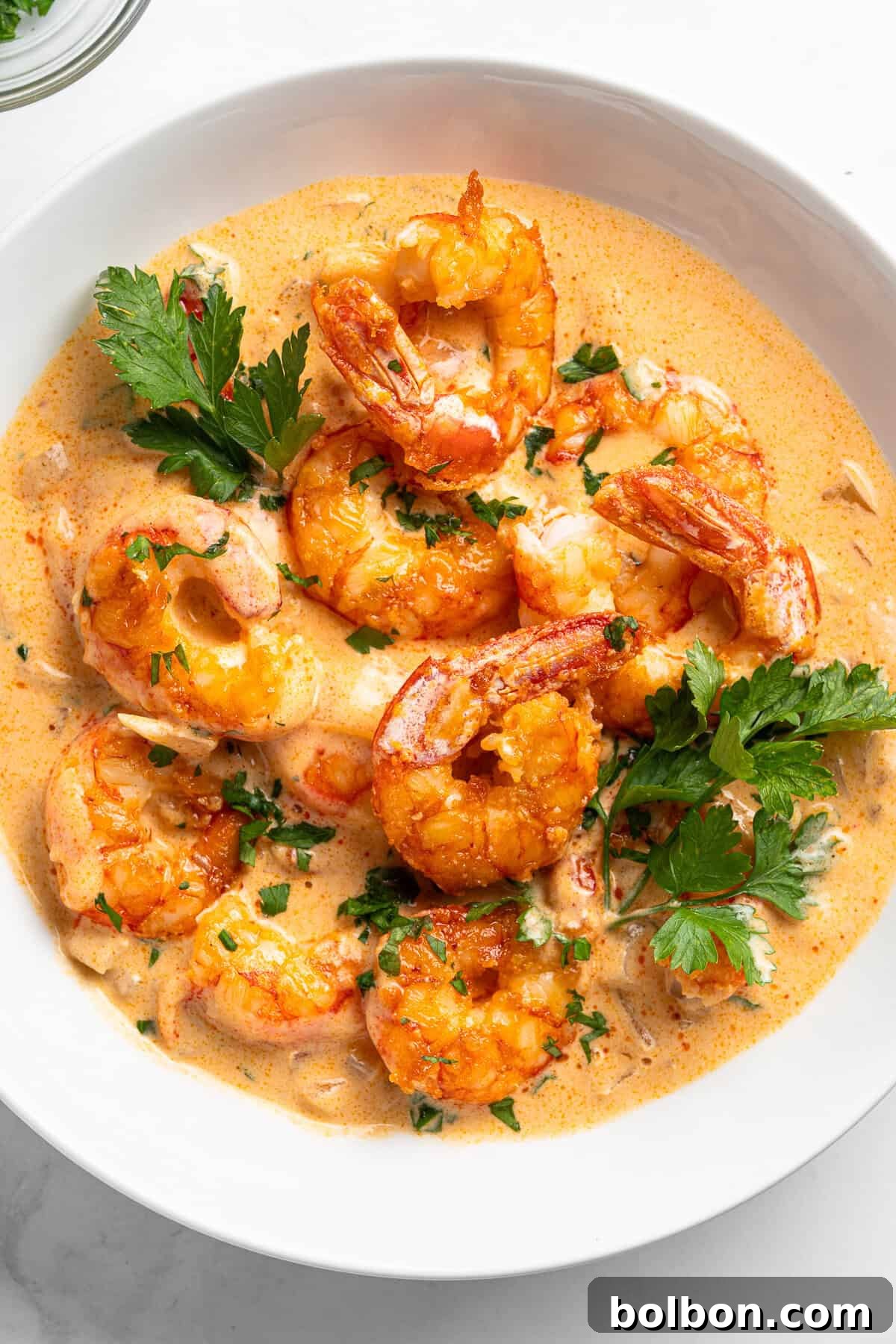 A pristine white bowl filled with creamy garlic shrimp, beautifully garnished with fresh parsley. The shrimp, cooked to a perfect golden hue, are nestled within a rich, orange-tinted sauce. The inviting dish is elegantly presented against a light background.