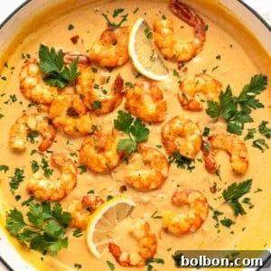 A pan filled with creamy orange sauce topped with cooked shrimp, garnished with fresh parsley and lemon slices. The shrimp are arranged attractively on the sauce, creating a visually appealing dish. Lemon wedges are placed on the side.