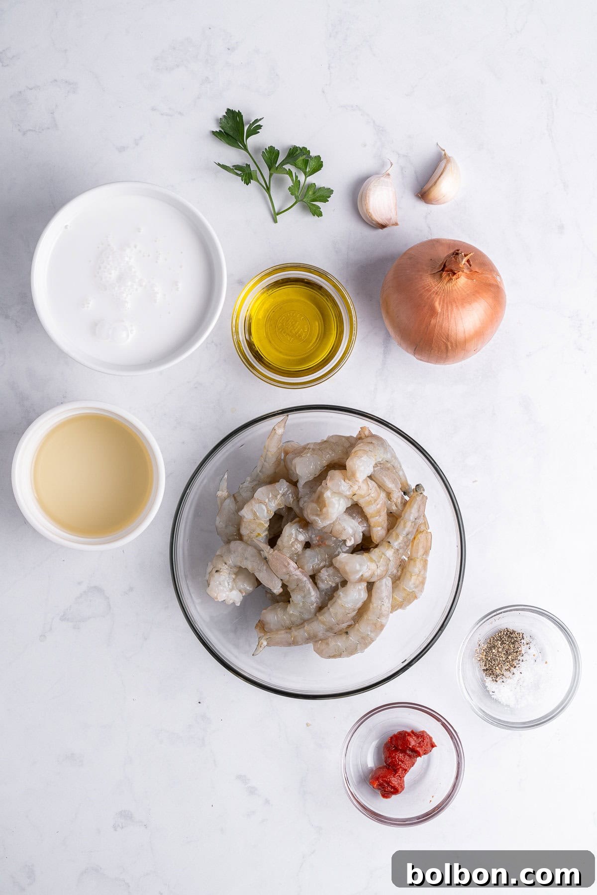 Top-down view of cooking ingredients on a white surface, including raw seafood in a bowl, an onion, garlic cloves, olive oil, coconut milk, white wine, tomato paste, parsley, salt, and pepper, showcasing the fresh components ready for preparation.