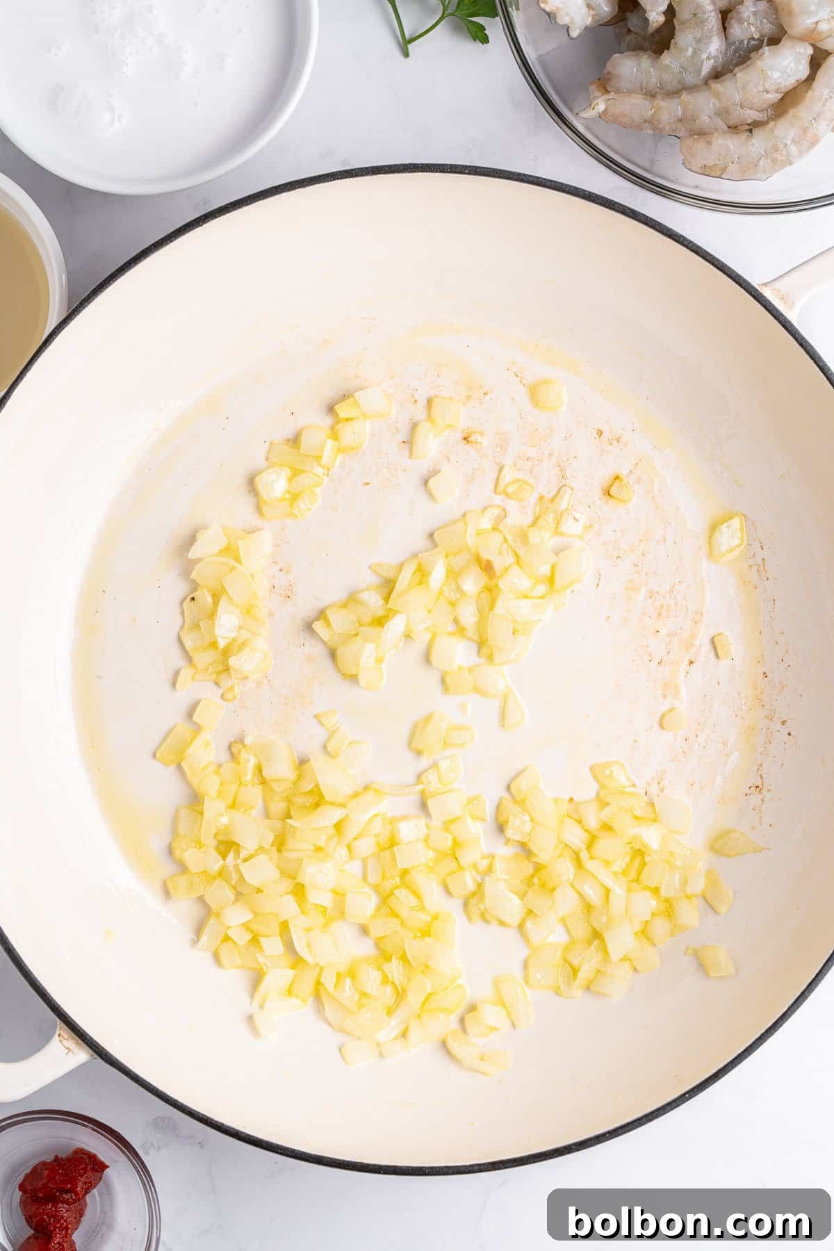 Finely chopped onions sautéing in a sleek white skillet, surrounded by complementary ingredients such as vibrant tomato paste and a small bowl of white liquid, all set on a pristine white countertop.