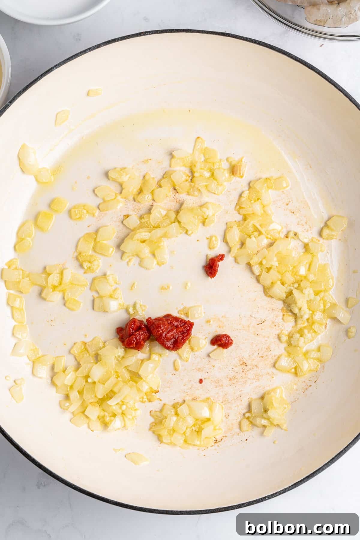 A harmonious blend of chopped onions and minced garlic gently sautéing in a white pan, with a dollop of rich tomato paste nestled in the center. The ingredients are lightly cooked and glisten with olive oil, all resting on a elegant white marble surface.