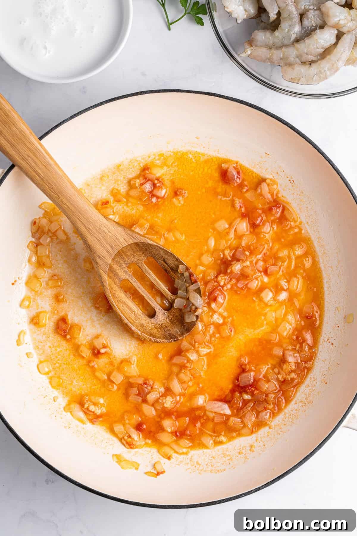 A pan with perfectly cooked onions and diced tomatoes suspended in a rich, reddish oil mixture, stirred gently by a sturdy wooden spoon. Nearby, a small bowl holds a delicate white sauce, all beautifully arranged on a white marble surface.