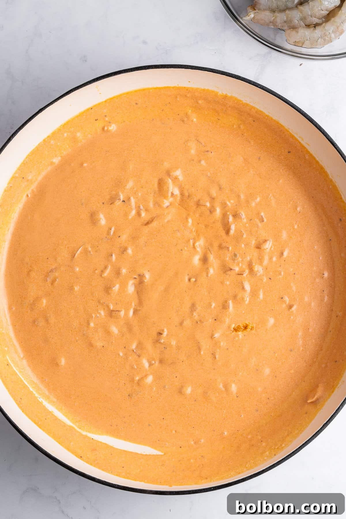 A pan, brimming with creamy, textured sauce, rests gracefully on a pristine white marble surface. In the upper corner, a bowl containing fresh, raw shrimp is partially visible, hinting at the culinary journey ahead.