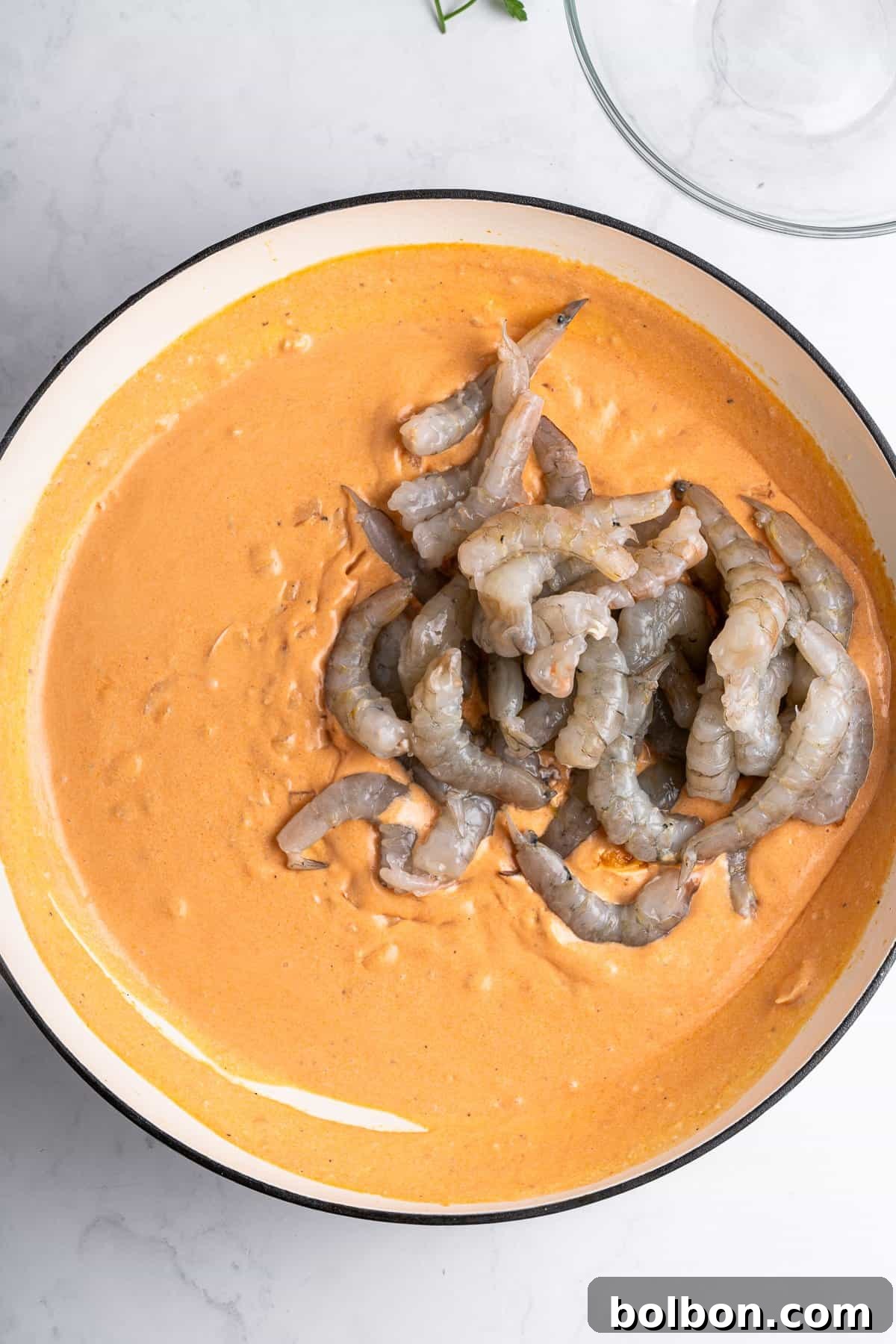 A skillet filled with a smooth, rich creamy sauce, topped with uncooked shrimp, creating a beautiful contrast. The skillet sits on a white marble surface, with a glass bowl partially visible in the corner.